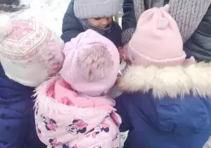 zieci stoją wok&oacute;ł nauczycielki. Pochylają się. Wybierają ziarna z torebki.