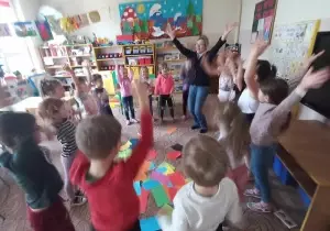 Dzieci stoją w kole. W kole leżą kolorowe kartki. Dzieci podnoszą ręce do g&oacute;ry.