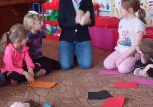 Pani psycholog siedzi na dywanie w kole. Przed nią leżą kolorowe kartki. Obok niej siedzą dzieci.