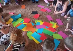 Dzieci z panią psycholog siedzą w kole. Na środku leżą kolorowe kartki.
