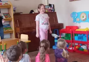 Dziewczynka stoi na środku. Inne dzieci siedzą na dywanie. Patrzą się na nią.