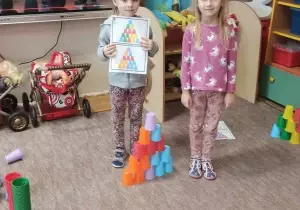 Dwie dziewczynki stoją na dywanie. Jedna znich trzyma kartkę z wydrukowanymi kolorowymi karteczkami.Między nimi stoi nudowla z kolorowych kubeczk&oacute;w.