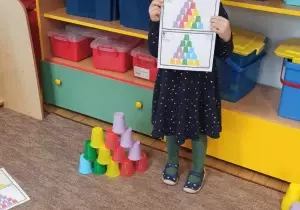 Dziewczynka stoi na dywanie w klasie. Trzyma w rękach kartkę z wydrukowanymi kolorowymi kubkami. Obok niej stoi budowla z kubeczk&oacute;w.
