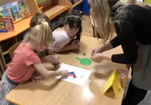 Nauczycielka stoi przy stoliku. Dzieci siedzą. Nauczycielka pomaga dzieciom ułożyć puzzle.