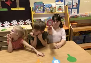 Tr&oacute;jka dzieci siedzi przy stoliku. Dziewczynka podnosi cyferkę 2 naklejoną na czerwonym kartoniku.