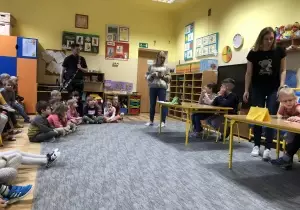 Tr&oacute;jka dzieci siedzi przy stoliku. Nauczyciel stoi przy stoliku. Drugi nauczyciel stoi z gitarą. Pozostałe dzieci siedzą na krzesełkach jako widownia.