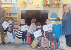 Dzieci stoją w sali przedszkolnej. Obok nich stoi leśnik i nauczycielka. Dzieci trzymają w rękach dyplomy i nagrody. Uśmiechają się do zdjęcia.