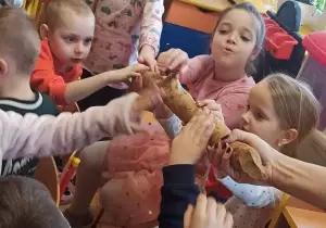 Dzieci siedzą na krzesełkach. Wyciągają ręce do przodu i dotykają kawałek drewna obgryzionego przez bobry.