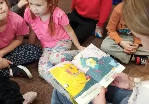 Dzieci siedzą po turecku na dywanie. Absolwentka trzyma w ręku książkę. Czyta dzieciom.