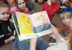Dzieci siedzą po turecku na dywanie. Absolwentka trzyma w ręku książkę. Czyta dzieciom.