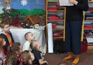 Pani stoi przed dziećmi. Dzieci siedzą na dywanie. Pani trzyma w ręku kartę pracy dla dzieci