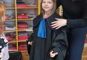 Dziewczynka stoi ubrana w togę. Za nią stoi pani.