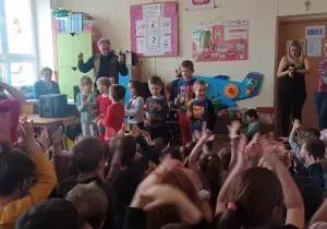Si&oacute;demka dzieci stoi i gra na r&oacute;żnych instrumentach. Za nimi siedzi pan grający na perkusji. Przed nimi dzieci, kt&oacute;re klaszczą, trzymają ręce w g&oacute;rze.