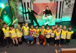 Dzieci stoją w teatrze. Są ubrane w kamizelki odbalskowe. Za nimi stoją aktorki. Aktorki trzymaja w rękach lalki bunraku