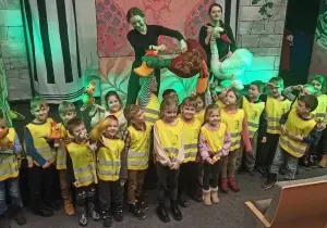 Dzieci stoją w teatrze. Są ubrane w kamizelki odbalskowe. Za nimi stoją aktorki. Aktorki trzymaja w rękach lalki bunraku