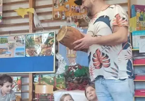 Pan stoi. Trzyma w ręku bęben. Obok niego siedzą dwiedziewczynki. Patrzą na niego. Przed nimi stoi bęben.