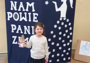 Dziewczynka stoi i trzyma w ręku bałwana. Uśmiecha się. Stoi na tle dekoracji co nam powie pani zima