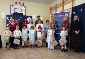 Grupowe zdjęcie uczestnik&oacute;w konkursu. Razem z nimi stoją nauczyciele i organizatorzy. Dzieci trzymają w rękach dyplomy i nagrody.