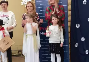 Stoi trzech nauczycieli. Przed nimi dwie dziewczynki i jeden chłopiec. Jedna z Pań bije brawo. Dzieci się uśmiechają. Jedna dziewczynka trzyma w ręku zabawkę bałwana. Druga klaszcze.