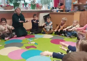 Dzieci siedzą na dywanie w klasie szkolnej. Razem z nimi siedzi nauczycielka. Dzieci wyciągają ręce do przodu.