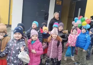 Dzieci stoją przed szkołą. Ubrane są w czapki i kurtki. Uśmiechają się do zdjęcia.