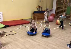Dzieci są na sali gimnastycznej. Jeżdżą na deskach.