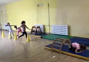 Dzieci wykonują zadania sportowe. Dwie dziewczynki biegną między pachołkami. Jedna dziewczynka czołga się pod drabinką na materacu.