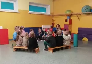 Dzieci siedzą na sali gimnastycznej w kręgu na ławkach. Wśr&oacute;d nich siedzi Pani. M&oacute;wi coś do nich.