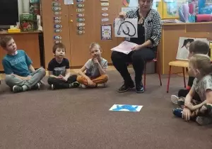 Pielęgniarka siedzi na krzesełku. Trzyma w ręku kartkę. Obok niej w kole siedzą dzieci. 