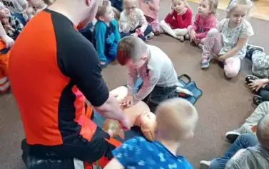 Ratownik jest w siadzie klęcznym. Przed nim leży fantom. Dziecko klęczące przed fantomem uciska fantoma na klatce piersiowej.