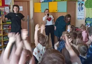 Dzieci trzymają ręce w g&oacute;rze. Klaszczą. Pani przed nimi tańczy.