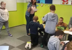 Nauczycielka trzyma w ręku pacynkę. Dzieci siedzą przy ławkach. Wycinają nożyczkami.