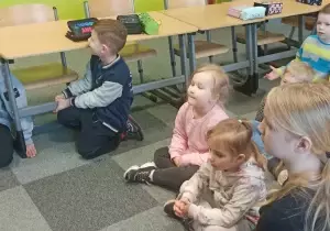 Dzieci siedzą na dywanie. Wok&oacute;ł nich stoją ławki.