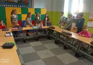 Uczniowie stoją przy ławkach w klasie szkolnej. Przed nimi siedzą przy ławkach dzieci z przedszkola.
