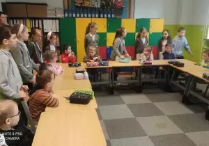 Uczniowie stoją przy ławkach w klasie szkolnej. Przed nimi siedzą przy ławkach dzieci z przedszkola.