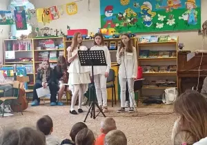 Na środku sali stoją trzy dziewczyny. Śpiewają piosenkę. Przed nimi stoi stojak z nutami. Siedzą r&oacute;wnież przedszkolaki, widownia.