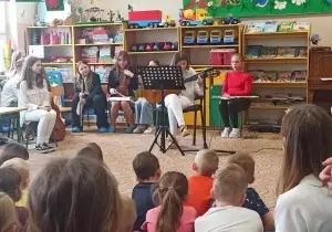 Dziewczynka siedzi na środku sali. Gra na gitarze. Przed nią stoi stojak z nutami. Siedzą r&oacute;wnież przedszkolaki, widownia.