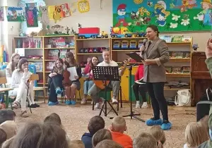 Dziewczynka siedzi na środku sali. Trzyma gitarę. Przed nią stoi stojak z nutami. Siedzą r&oacute;wnież przedszkolaki, widownia. Obok stoi prowadząca występ. Trzyma w rękach mikrofon i kartkę z tekstem z kt&oacute;rej czyta.