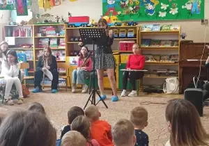 Dziewczynka stoi na środku sali. Gra na skrzypcach. Przed nią stoi stojak z nutami. Siedzą r&oacute;wnież przedszkolaki, widownia.