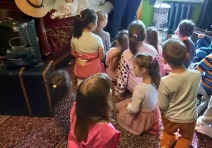 Dzieci siedzą na dywanie. Kustosz pokazuje im mały piecyk. Obok nich znajduje się starodawne, drewniane ł&oacute;żko. Na nim jest pościel i lalka.