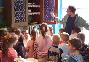 Dzieci stoją przed eksponatem, starodawnym kredensem. Pan wskazuje ręką na kredens. Opowiada o nim.