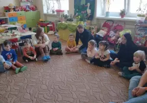 Dzieci wraz z rodziacmi siedzą na dywanie.