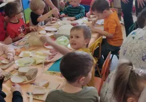Dzieci siedzą przy stolikach. Rodzice stoją przy dzieciach. Razem wykonują kanapki.
