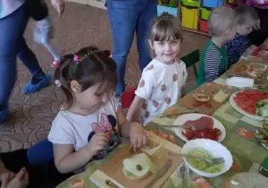Dwie dziewczynki siedza przy stoliku. Przed nimi stoją miseczki z produktami do robienia kanapek. Dziewczynka ozdabia kanapkę.