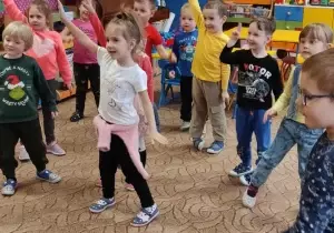 Dzieci stoją w rozsypce. Pokazują gest. Jedna ręka do g&oacute;ry w bok.