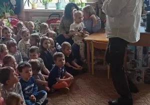 Pan Witek m&oacute;wi przez mikrofon. Dzieci siedzą na dywanie. Słuchają.
