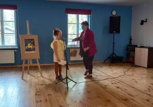 Dziewczynka stoi przy mikrofonie. Obok stoi Pani prowadząca konkurs. Niesie w ręku koszyk.