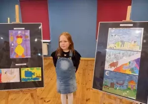 Julia stoi między dwiema sztalugami. Na nich są prace dla dzieci. Uśmiecha się do zdjęcia