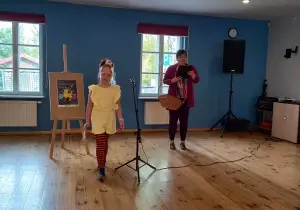 Dziewczynka schodzi ze sceny po recytacji wiersza. Pani prowadząca konkurs m&oacute;wi przez mikrofon. Trzyma w ręku koszyk i kartkę.