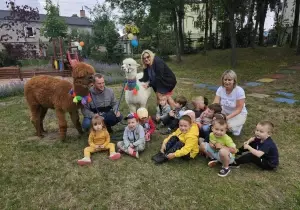Dzieci siedzą na trawie. Z tyłu stoją dwie alpaki. Brązowa i biała. Z tyłu nauczycielka przytula się do alpaki białej.